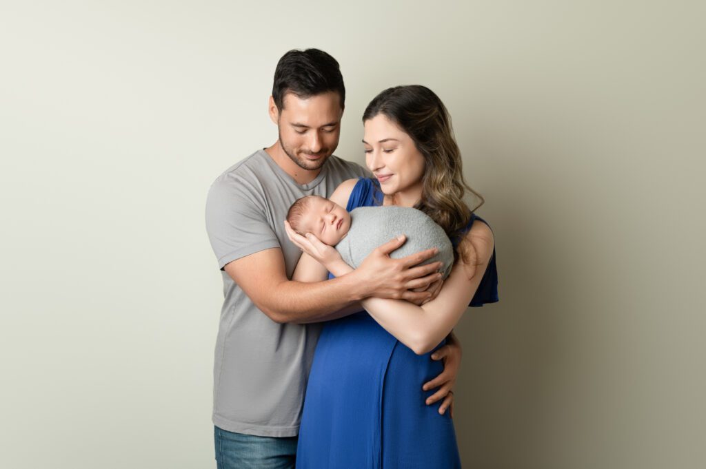 Parents standing close together, lovingly holding their newborn wrapped in gray during a home session.