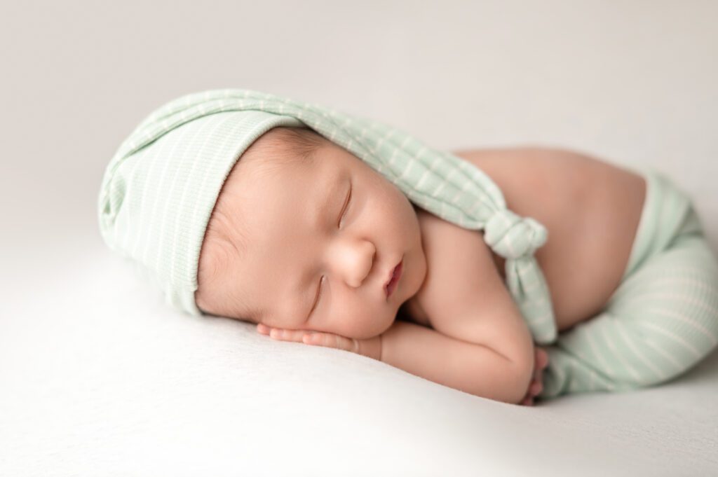 Newborn in soft green striped pajamas and matching sleepy cap, resting on a white blanket.