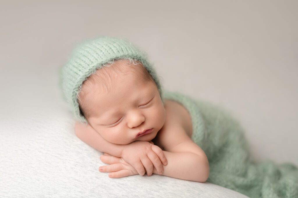 Baby sleeping on a textured white surface, wrapped in a mint green knitted wrap and bonnet.