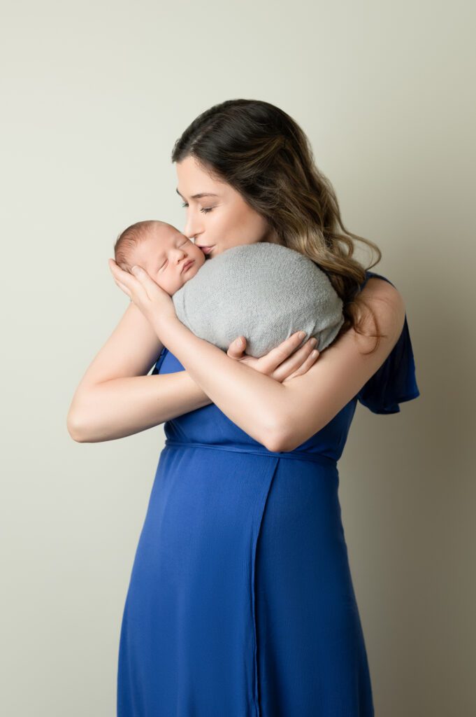 Mother holding her swaddled newborn close, dressed in a royal blue gown during an in-home photo session.