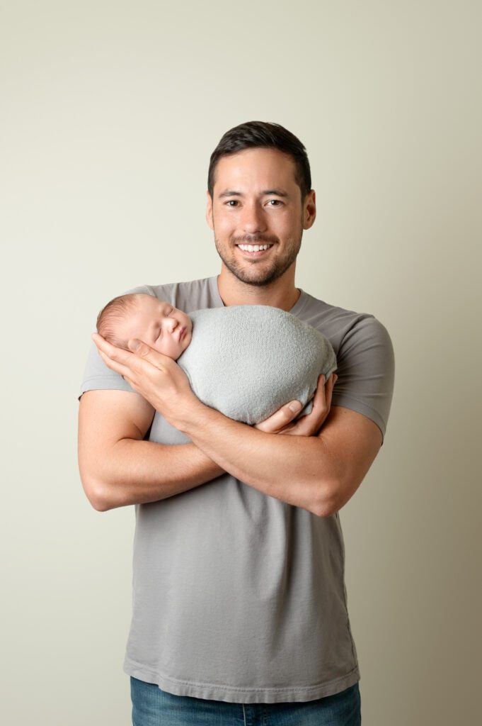 Father smiling while cradling his newborn in a gray swaddle against a neutral backdrop.


