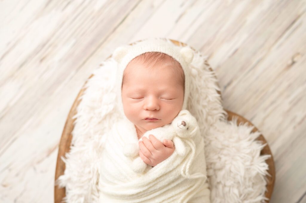 Newborn swaddled in white, wearing a bear-ear bonnet, gently holding a tiny teddy bear.