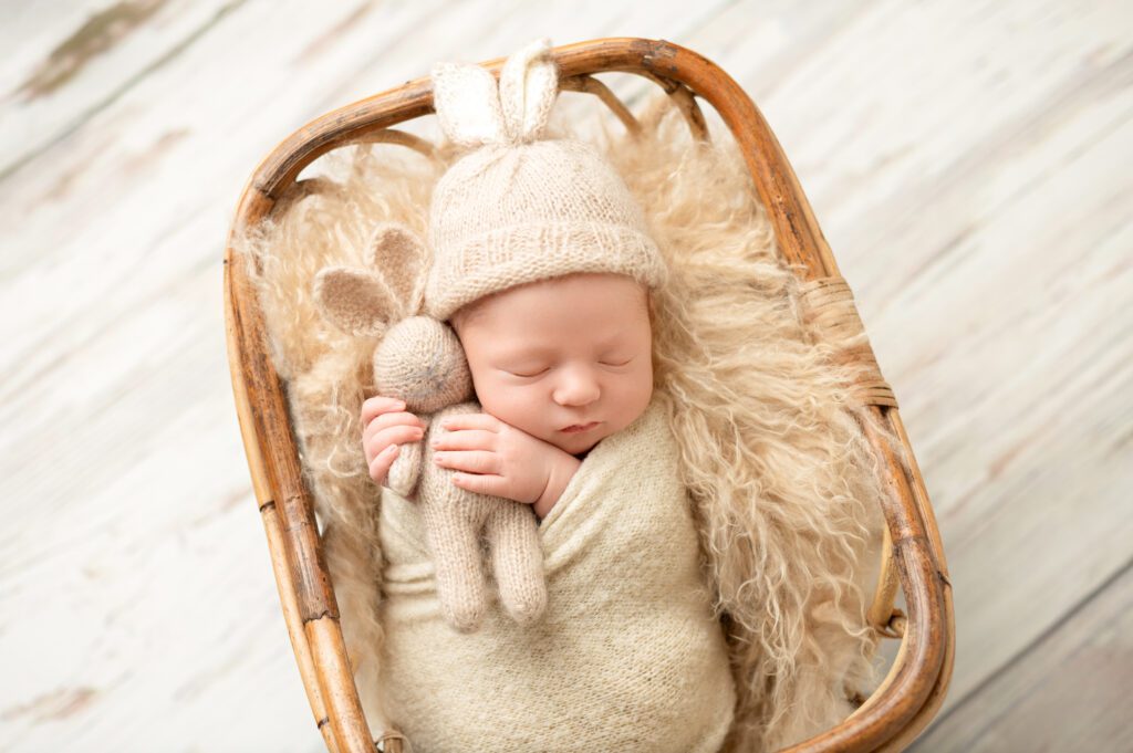 Baby in a beige wrap and knit bunny hat, sleeping peacefully in a wicker basket with soft textures.
