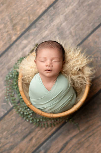 Newborn wrapped in soft green swaddle, sleeping peacefully in round basket.