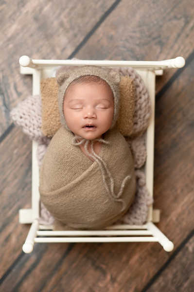 Newborn dressed in cozy bear outfit wrapped in brown blanket, posed in small crib.