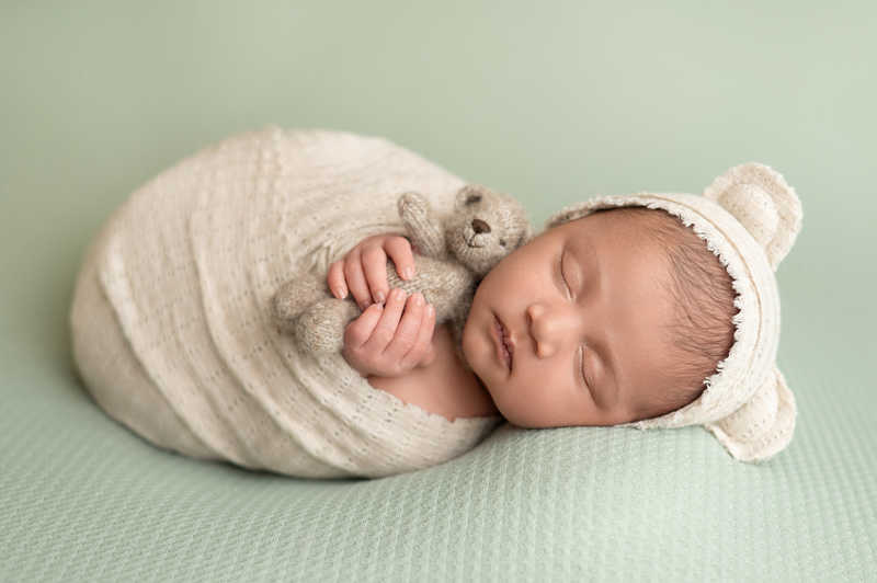 Newborn baby wrapped in cream blanket with bear bonnet and small stuffed toy.