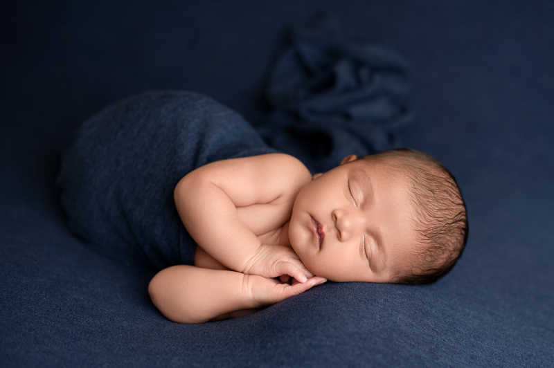 Newborn boy in dark blue wrap sleeping on his side during at-home photo session.