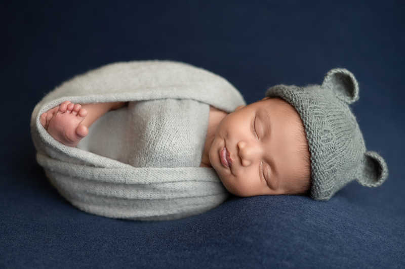 Newborn baby peacefully sleeping wrapped in gray blanket with tiny bear hat.